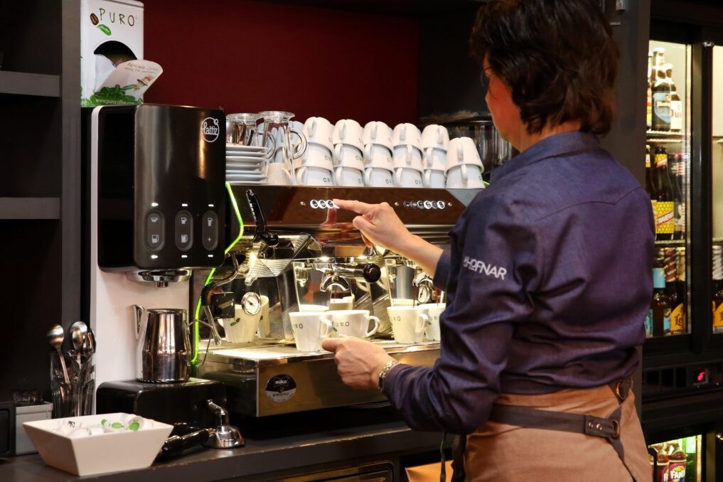 Horecamedewerker in een grijsblauw bloesje zet bij een grote koffiemachine een aantal kopjes koffie.