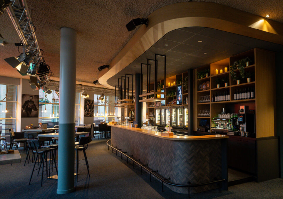 Leeg theatercafé met de bar prominent in het zicht. Bar die donkere houten uitstraling heeft met rondingen aan de uiteindes.